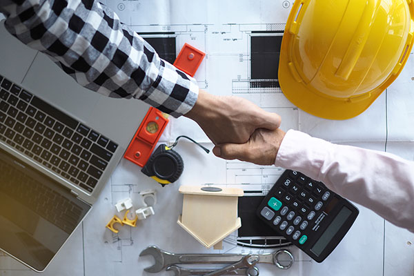Two people shake hands over a desk with tools and a hard hat on it